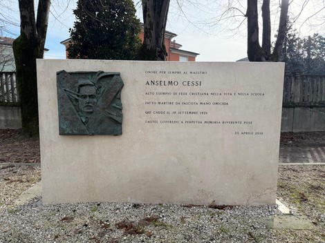 Monumento commemorativo a Anselmo Cessi, con un rilievo e iscrizioni, circondato da alberi.