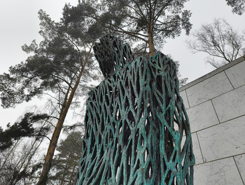 Scultura in bronzo al cimitero militare italiano di Zehlendorf, circondata da alberi. Atmosfera di riflessione.