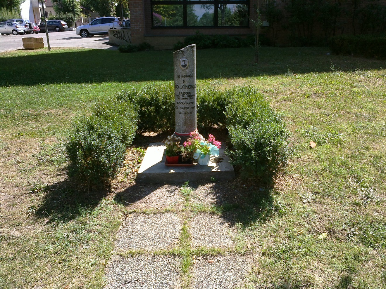 Cippo commemorativo circondato da arbusti e fiori, inserito in un contesto verde e curato.