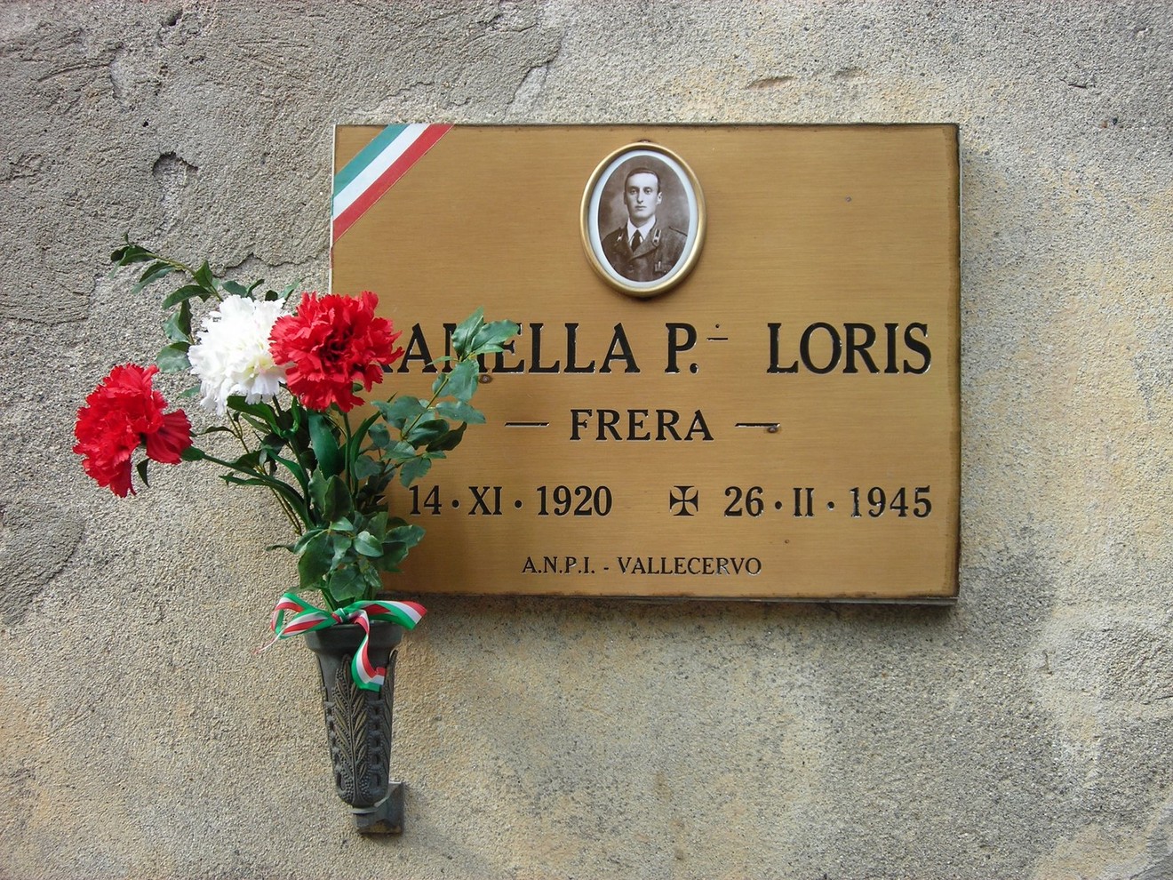 Targa commemorativa dedicata a Loris Ramella, con fiori e simboli tricolori, in memoria del suo sacrificio.