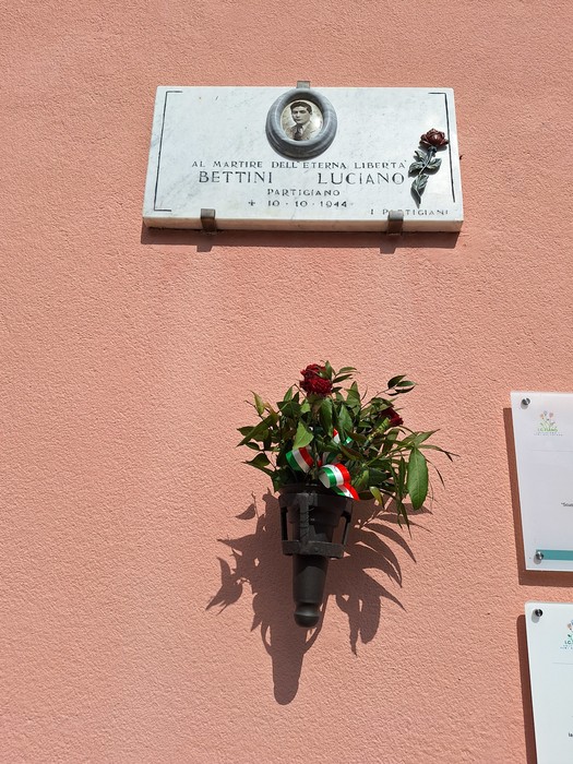 Lapide commemorativa a Luciano Bettini, partigiano, decorata con fiori e simboli patriottici.