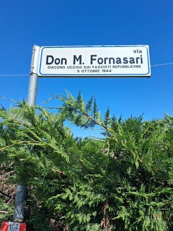 Cippo commemorativo dedicato a Don M. Fornasari, con sfondo di cielo blu.