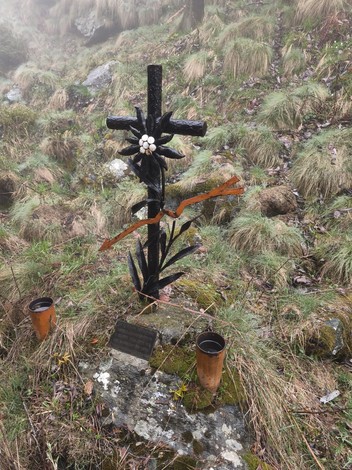 Croce in metallo nero con stella alpina, targa e bicchieri votivi, su base in pietra nel bosco.