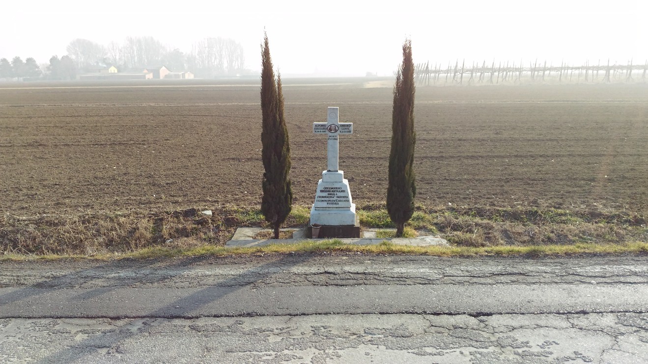 Croce commemorativa con cipressi, circondata da un vasto campo arato e un paesaggio rurale.