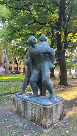 Scultura in bronzo raffigurante due figure umane che si abbracciano, immersa in un verde rigoglioso.