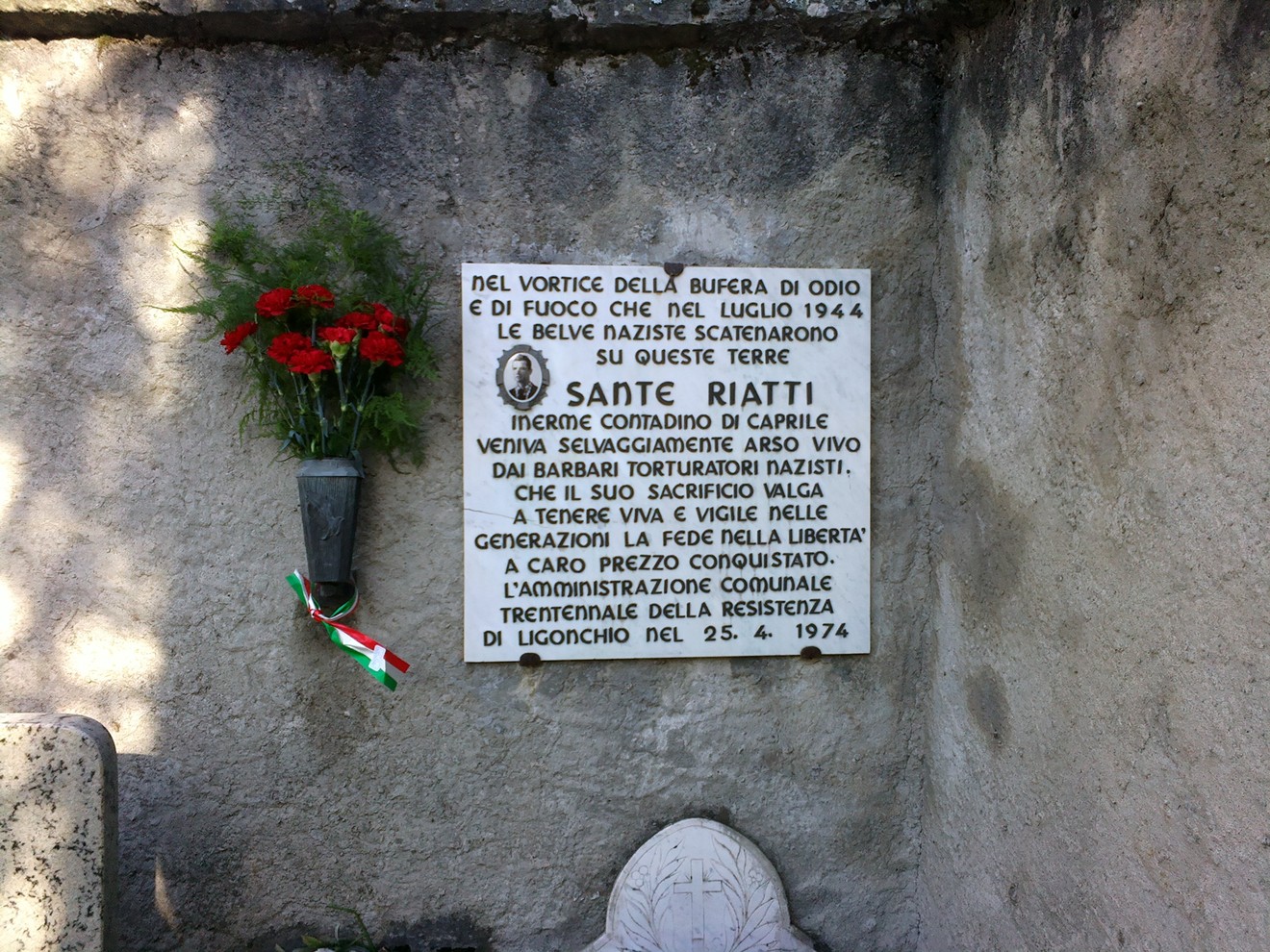 Lapide commemorativa a Riatti in memoria delle vittime dei nazisti, con fiori e bandiera italiana.