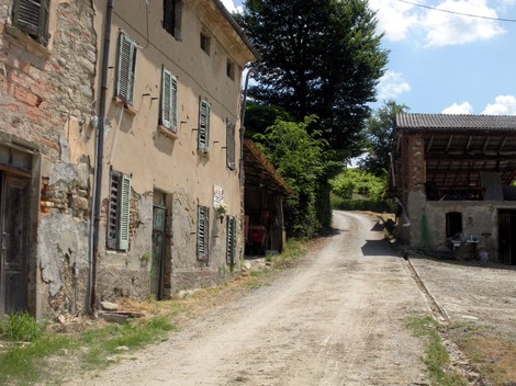 Strada sterrata che conduce a un'area rurale, con edifici in rovina e verde intorno. Atmosfera di abbandono.