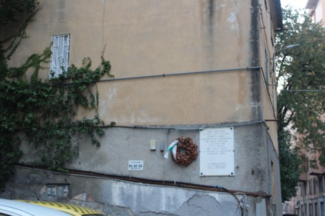 Lapide commemorativa su un muro, circondata da vegetazione, con una corona di fiori appesa.