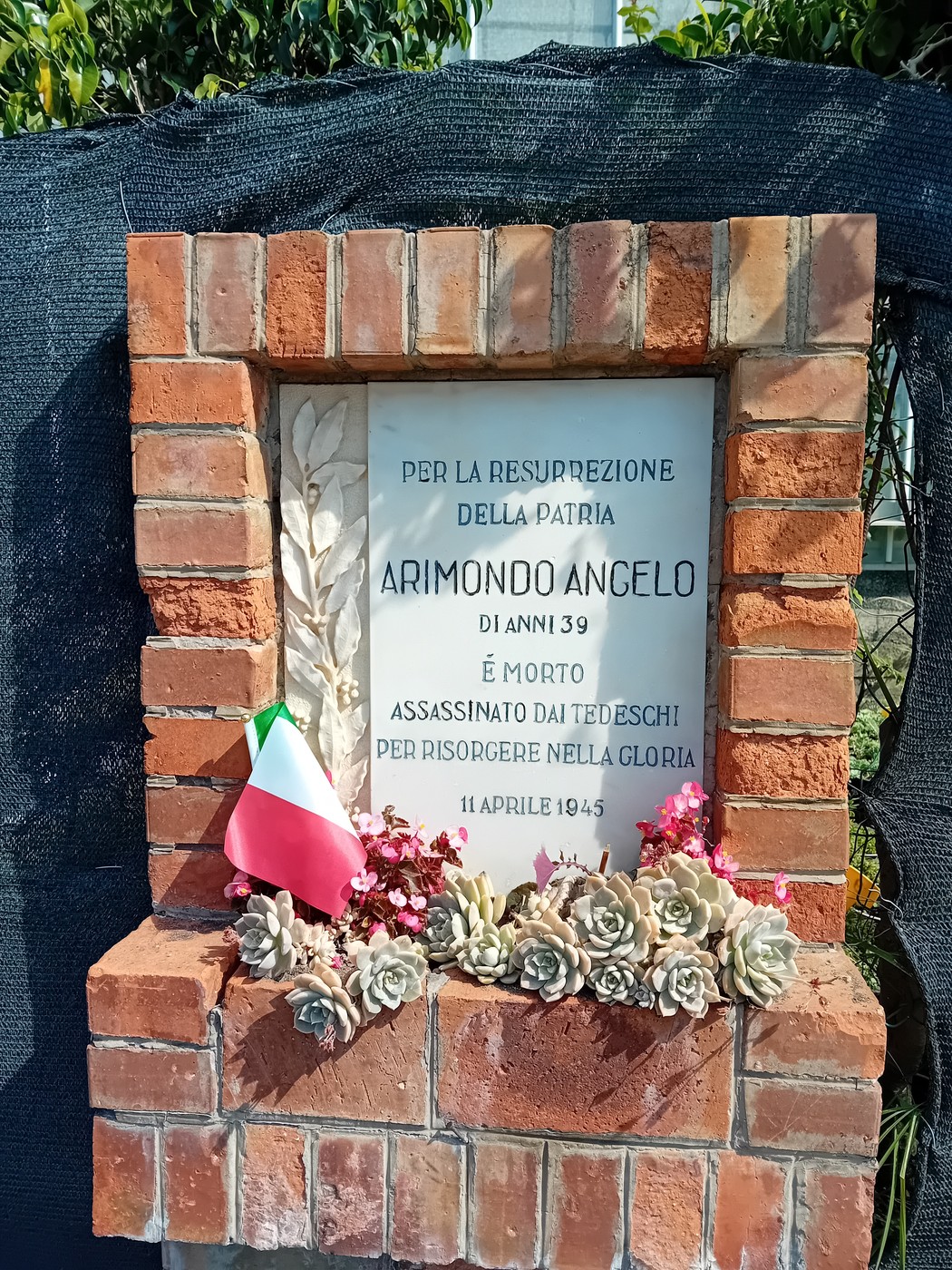 Lapide commemorativa dedicata ad Arimondo Angelo, con decorazioni floreali e il tricolore italiano.