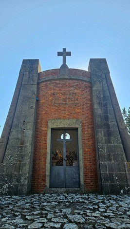 Cappella in pietra e mattoni con croce sulla facciata, portone in ferro e iscrizione sopra l'ingresso.