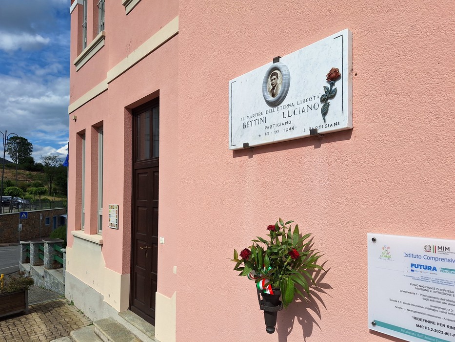 Lapide commemorativa dedicata a Luciano Bettini, posta su un edificio con un fiore collocato in onore della sua memoria.