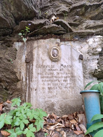 Lapide commemorativa di Mario Canavese, situata tra la vegetazione, con un'immagine e iscrizioni sulla pietra.