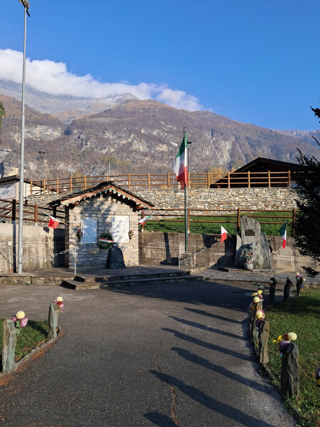 Piccolo monumento in pietra con lapide e iscrizione, bandiera italiana e recinto di legno sullo sfondo montano.