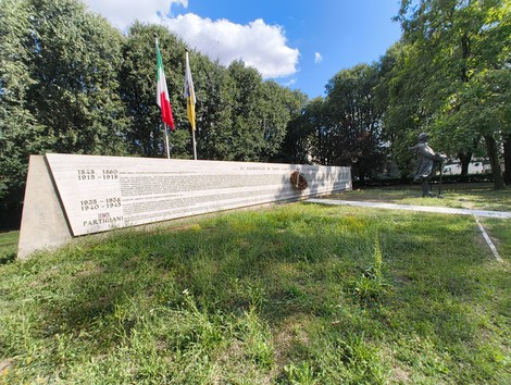 Monumento ai Caduti circondato da alberi, con bandiere italiane e simboli commemorativi visibili.