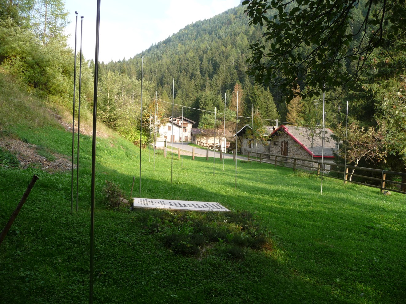Monumento ai Dodici Caduti immerso in un paesaggio verde, circondato da alberi e montagne.