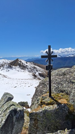 Croce metallica su una roccia, con vista panoramica di montagne innevate e cielo blu.