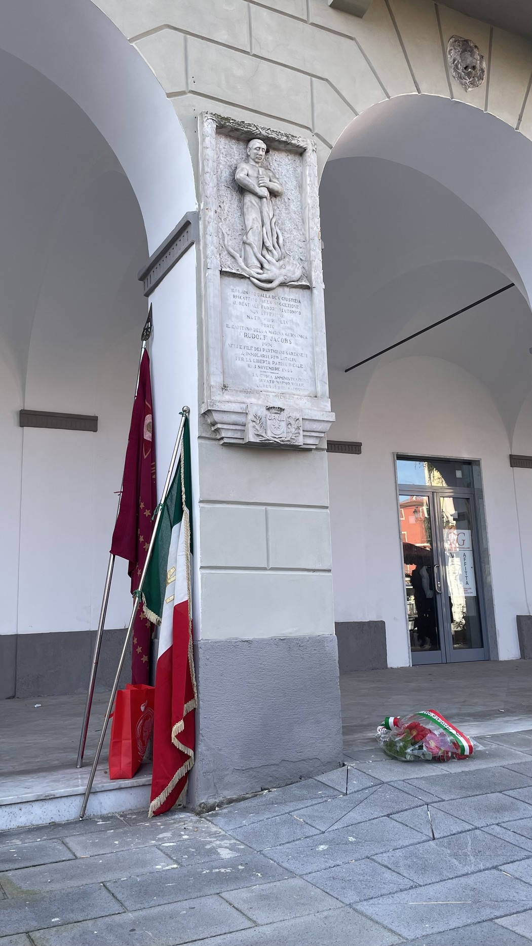 Lapide commemorativa dedicata a Jacobs, situata sotto un portico. Sullo sfondo, bandiere e un omaggio floreale.