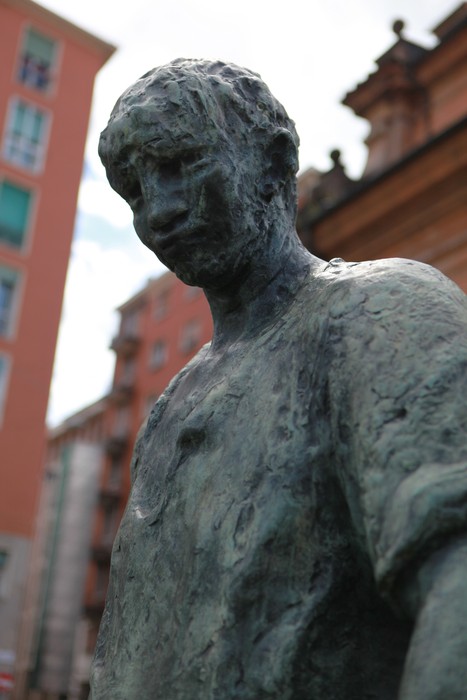 Scultura di un partigiano in bronzo, che trasmette un senso di forza e resistenza storica. Sullo sfondo edifici urbani.