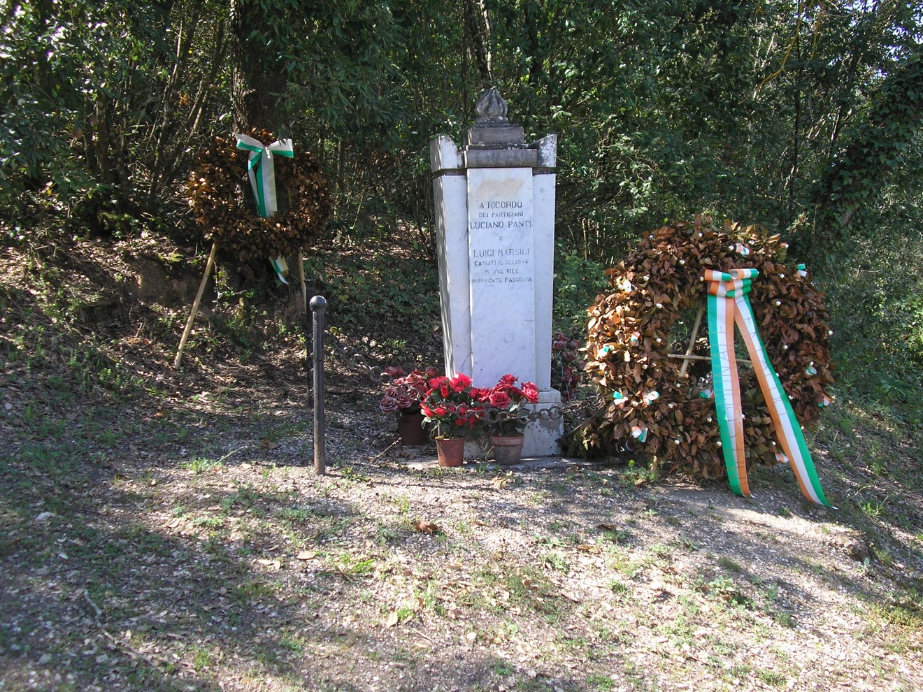 Cippo commemorativo ai caduti di Rigosecco, adornato con corone e fiori, immerso nella natura.