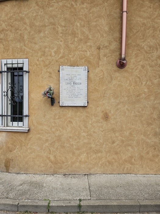 Lapide commemorativa su un muro di color ocra, con fiori e dedica a Ruggeri.
