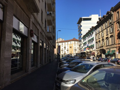 Strada cittadina con parcheggio, edifici in stile classico e moderno, cielo sereno e attrezzature commerciali visibili.