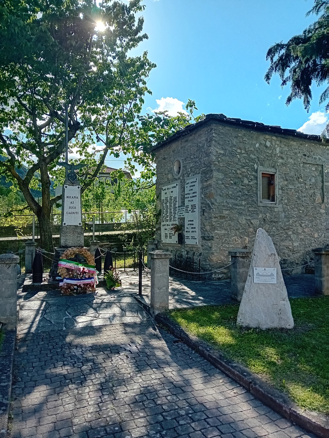 Monumento ai caduti con iscrizioni commemorative, circondato da alberi e un sentiero pavimentato.