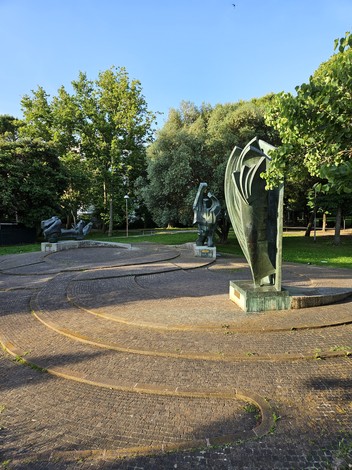 Monumento alla Resistenza, sculture in un parco con alberi e pavimentazione in mattoni. Atmosfera serena e verde.