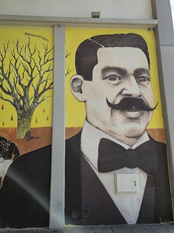 Murale colorato che ritrae un uomo con baffi, indossando un tailleur, sullo sfondo di un albero spoglio.