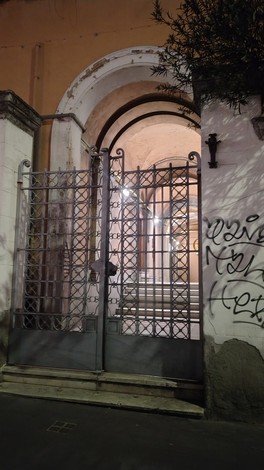 Porta in ferro battuto con cenni di graffiti, che conduce a un interno illuminato e architettonicamente interessante.
