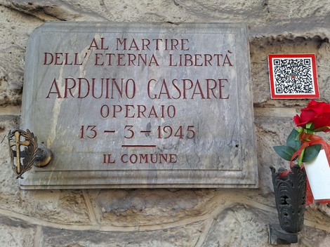 Lapide di marmo con iscrizione su parete in pietra, accanto una rosa rossa in un vaso e un codice QR.