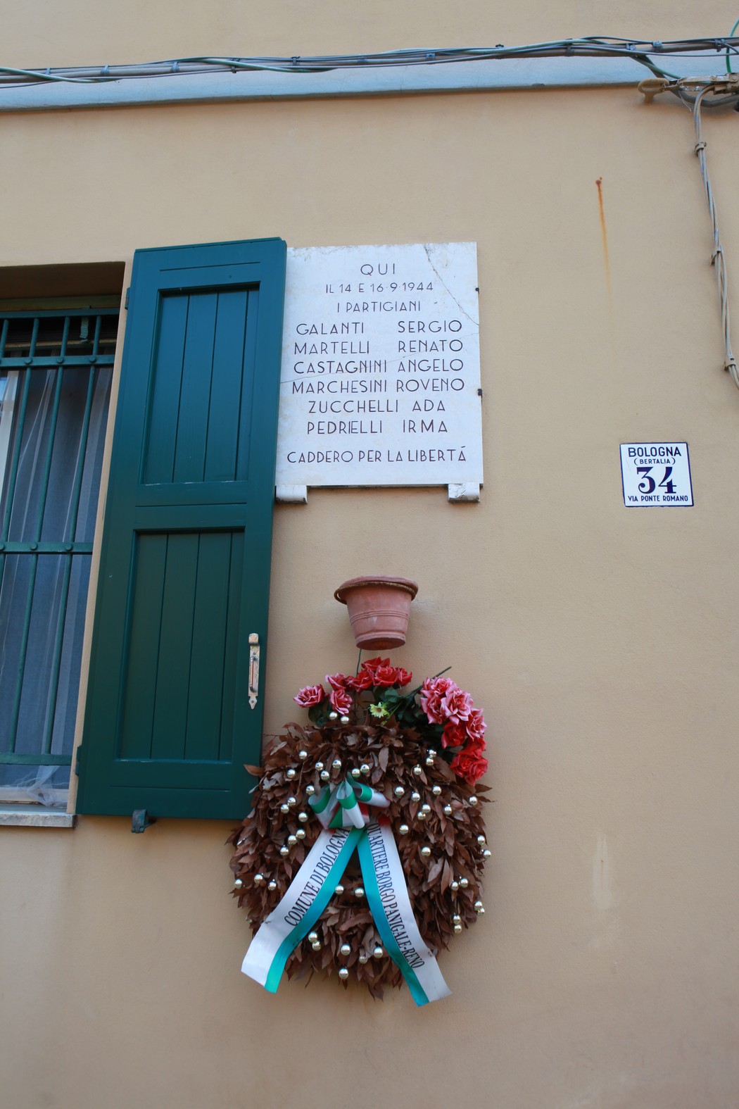 Lapide commemorativa con cerchio di fiori, dedicata ai caduti della 7° GAP, su un muro di un edificio.