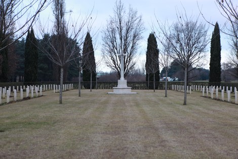 Cimitero di guerra del Commonwealth con croce centrale, alberi spogli e filari di lapidi. Atmosfera di solemnità e memoria.