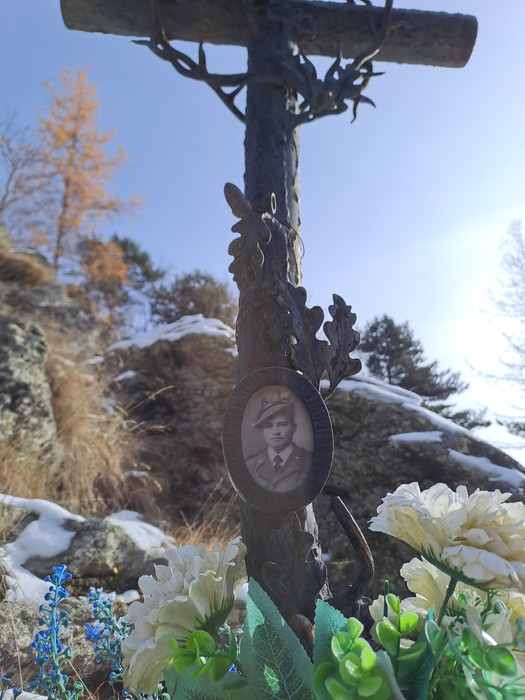 Croce in ferro battuto con medaglione fotografico di un uomo in divisa, tra fiori e rocce innevate.