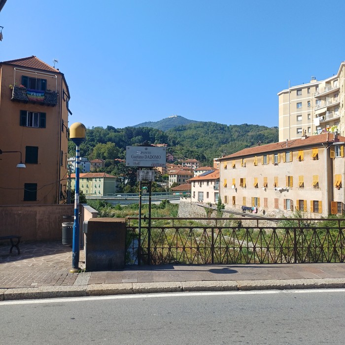Veduta urbana con targa stradale verticale su palo, edifici e colline sullo sfondo.