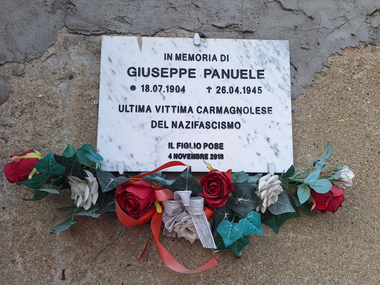 Lapide in marmo su parete intonacata, con iscrizione e corona di fiori artificiali appoggiata sotto.