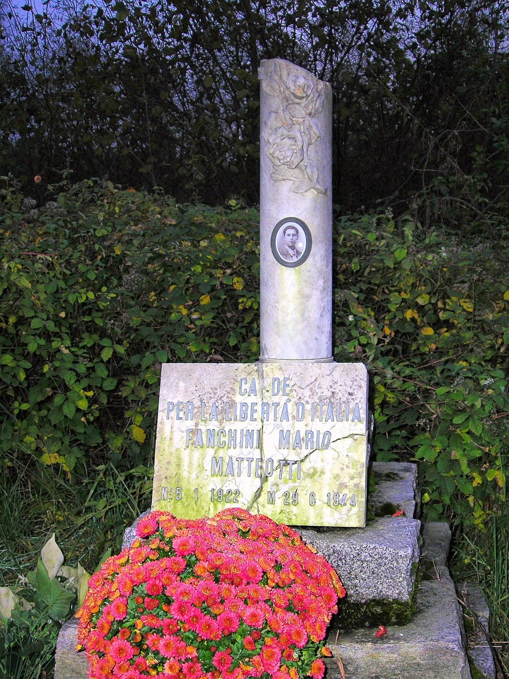Cippo commemorativo dedicato a Mario Fanchini, circondato da fiori in un contesto naturale.