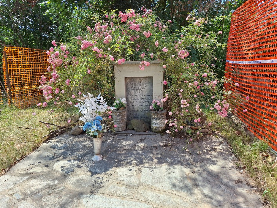 Cippo commemorativo circondato da fiori, con decorazioni floreali e un ambiente naturale sereno.