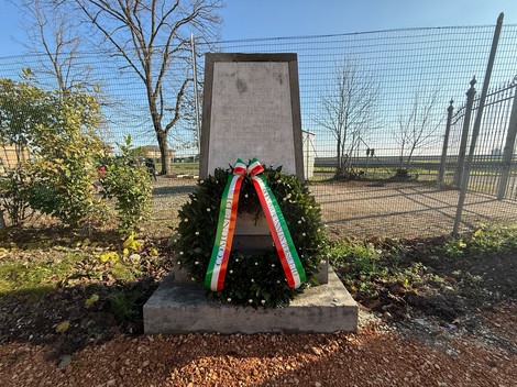 Monumento ai Caduti di Le Budrie con una corona di fiori, immerso in un'area verde e soleggiata.