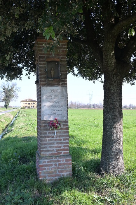 Cippo commemorativo affiancato da un albero in un campo verde, con una villa sullo sfondo.