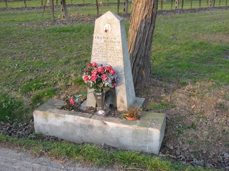 Cippo commemorativo a Masironi, con fiori e monumento in pietra, immerso nel verde della natura circostante.