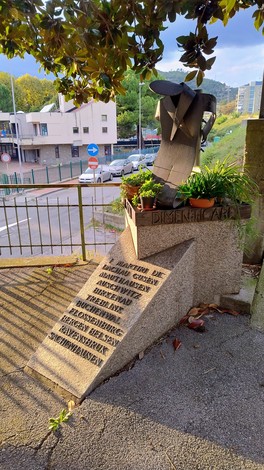 Monumento commemorativo ai deportati, con scultura e iscrizioni, immerso nel verde urbano.