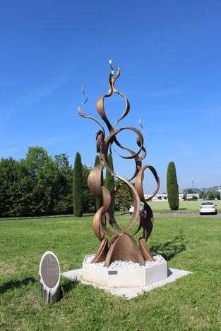 Monumento contemporaneo in metallo, con forme sinuose, immerso in un parco verde. Sullo sfondo, un cielo sereno.