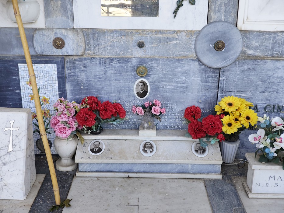 Loculi in marmo con ritratti fotografici, iscrizione e vasi di fiori artificiali colorati davanti.