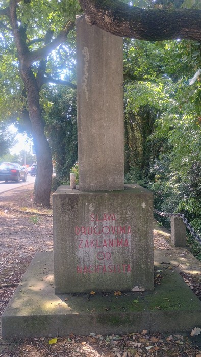 Monumento commemorativo con iscrizione, circondato da alberi e strada, in ricordo dei caduti per rappresaglia.