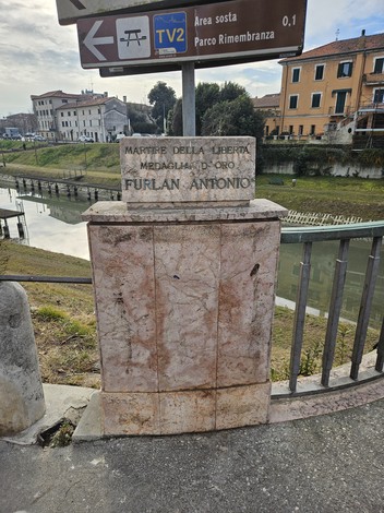Lapide commemorativa dedicata a Furlan Antonio, situata lungo il fiume in un contesto urbano.
