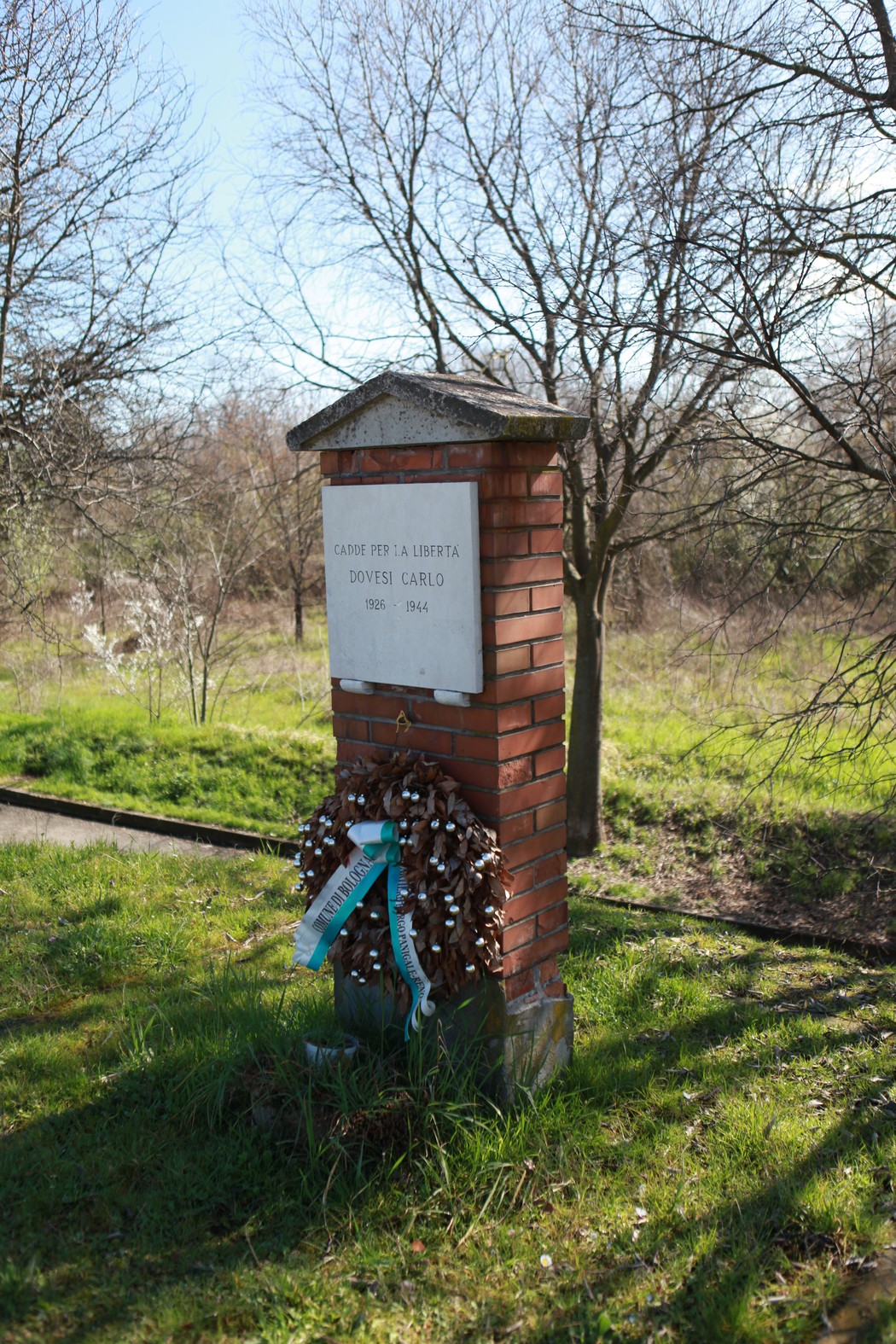Lapide commemorativa dedicata a Dovesi Carlo, circondata da vegetazione e alberi in un ambiente naturale.