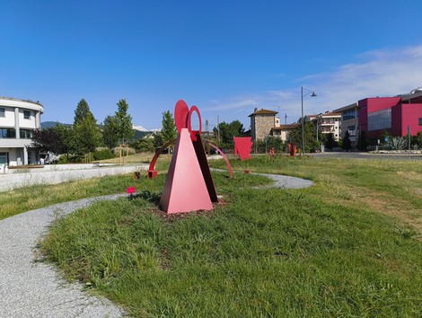 Scultura monumentale in un parco, con forme geometriche rosse su un prato verde e cielo sereno.