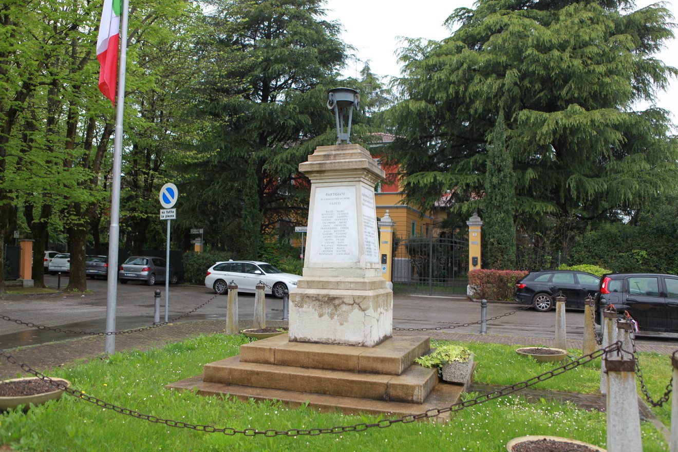 Monumento ai caduti per la Libertà, circondato da alberi e fiori, con bandiera italiana e lapidi commemorative.