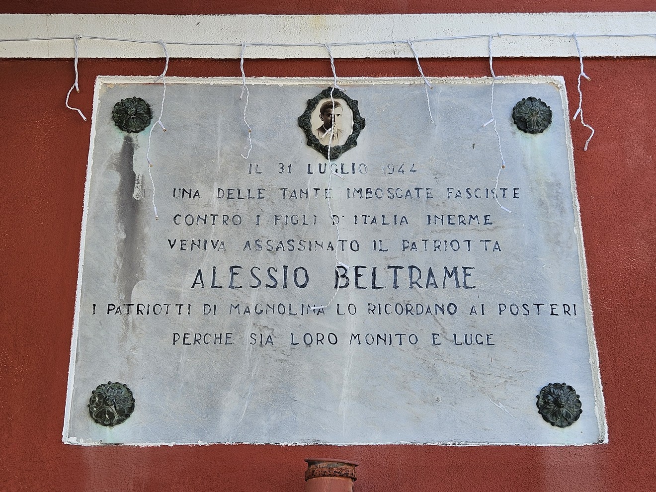 Lapide commemorativa dedicata ad Alessio Beltrame, patriota assassinato, con decorazioni e tono solenne.
