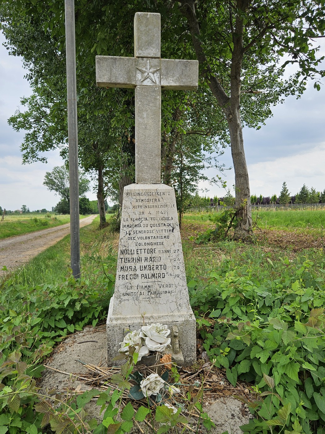 Cippo commemorativo con croce, circondato da vegetazione e ubicato lungo un sentiero.